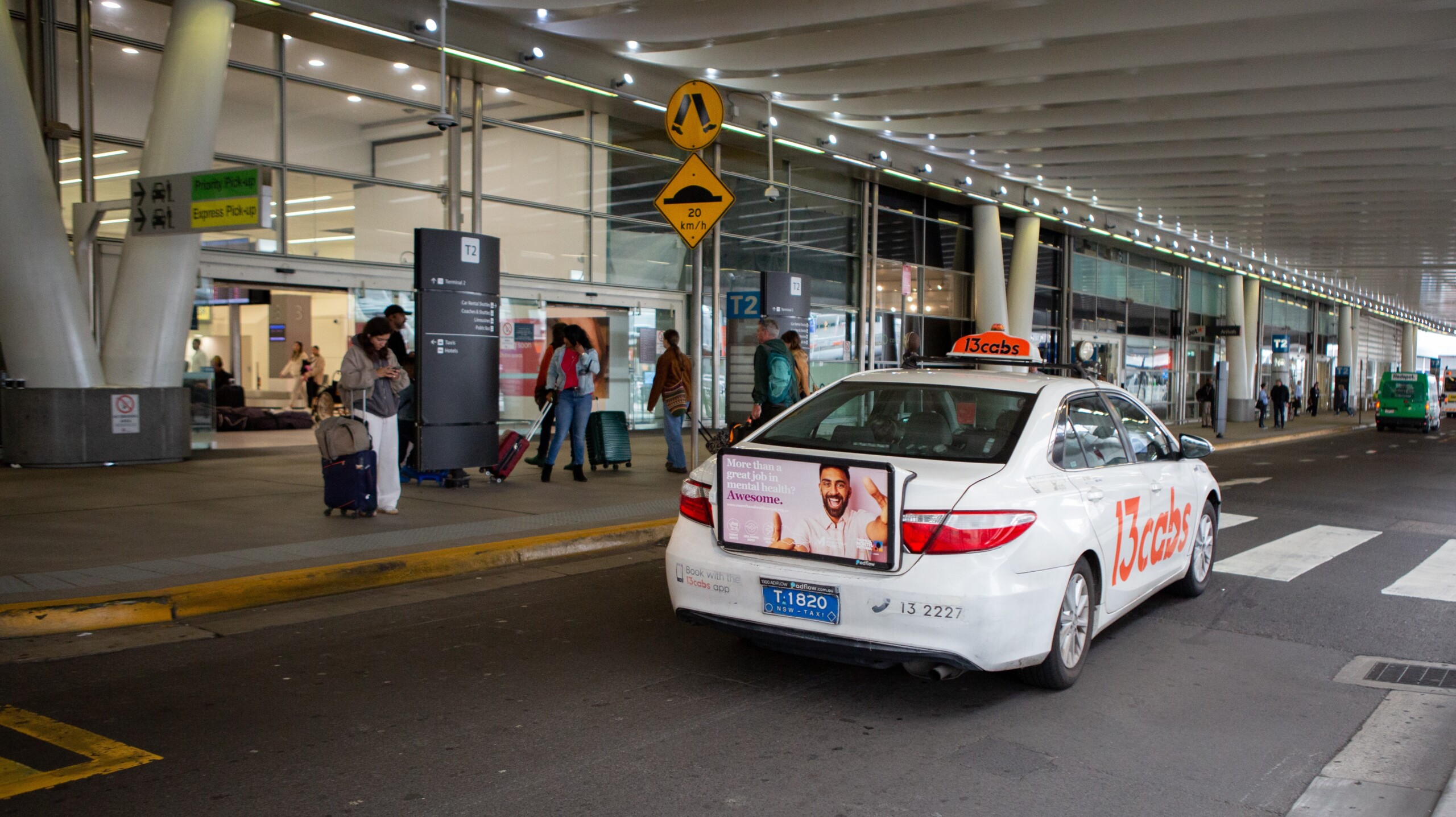 Taxi advertising displayed on the rear panel of a 13cabs taxi at Sydney Airport pick-up zone with travellers and terminal signage in the background.