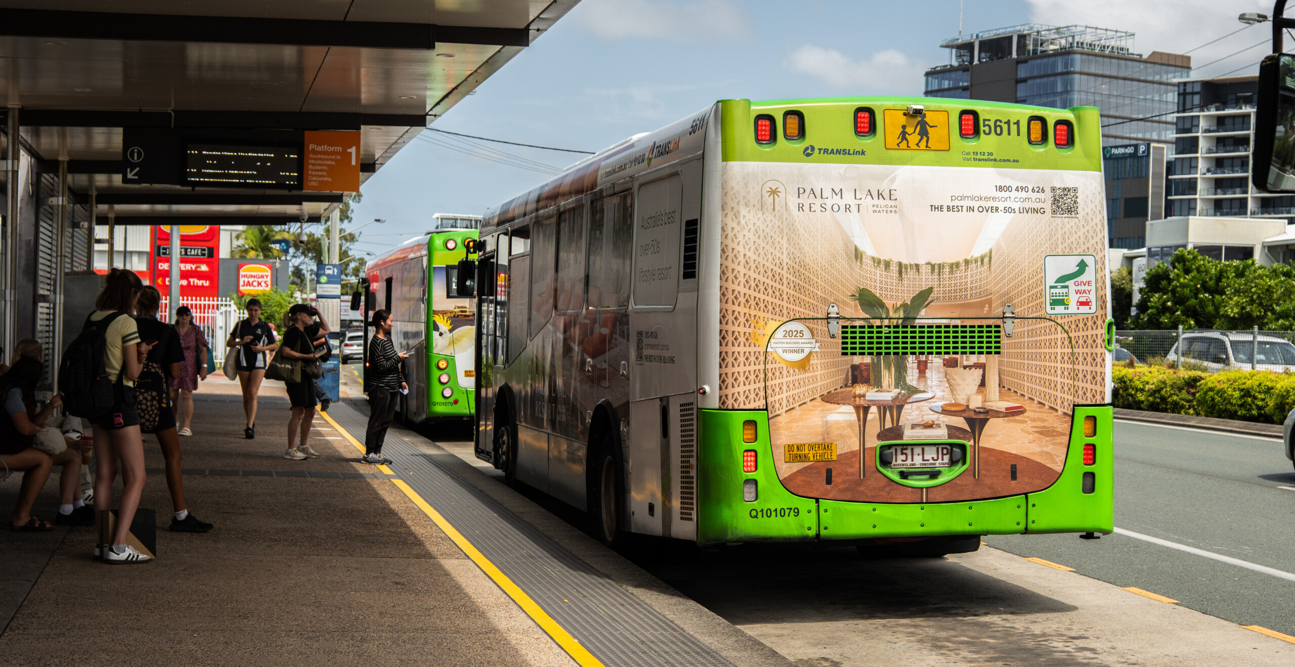 Bus featuring full-wrap transit advertising for Palm Lake Resort at a busy Sunshine Coast bus station, with passengers waiting and branded outdoor media displayed prominently.