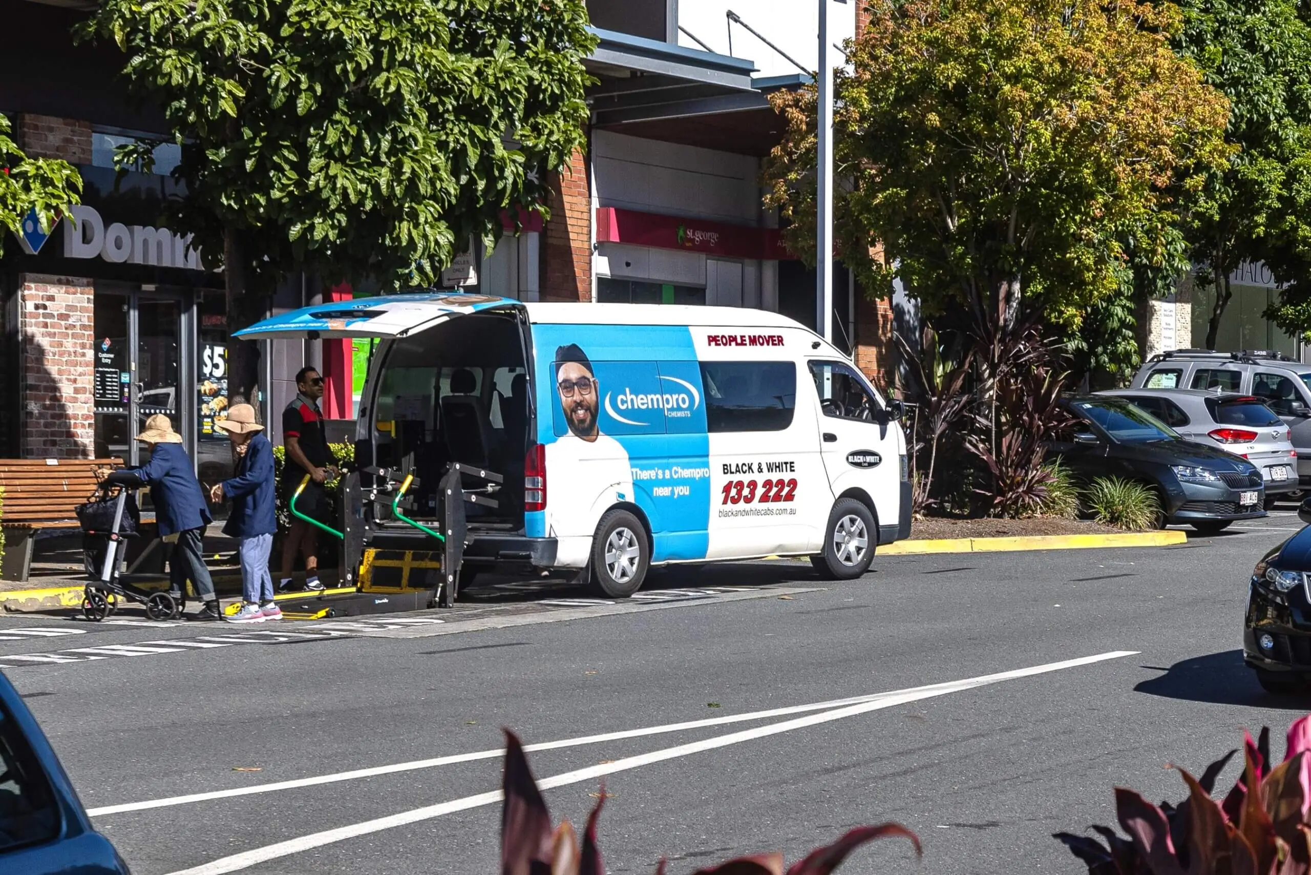Maxi taxi advertising on a Chempro-branded accessible people mover parked curbside, with passengers boarding via wheelchair ramp in a suburban shopping strip.