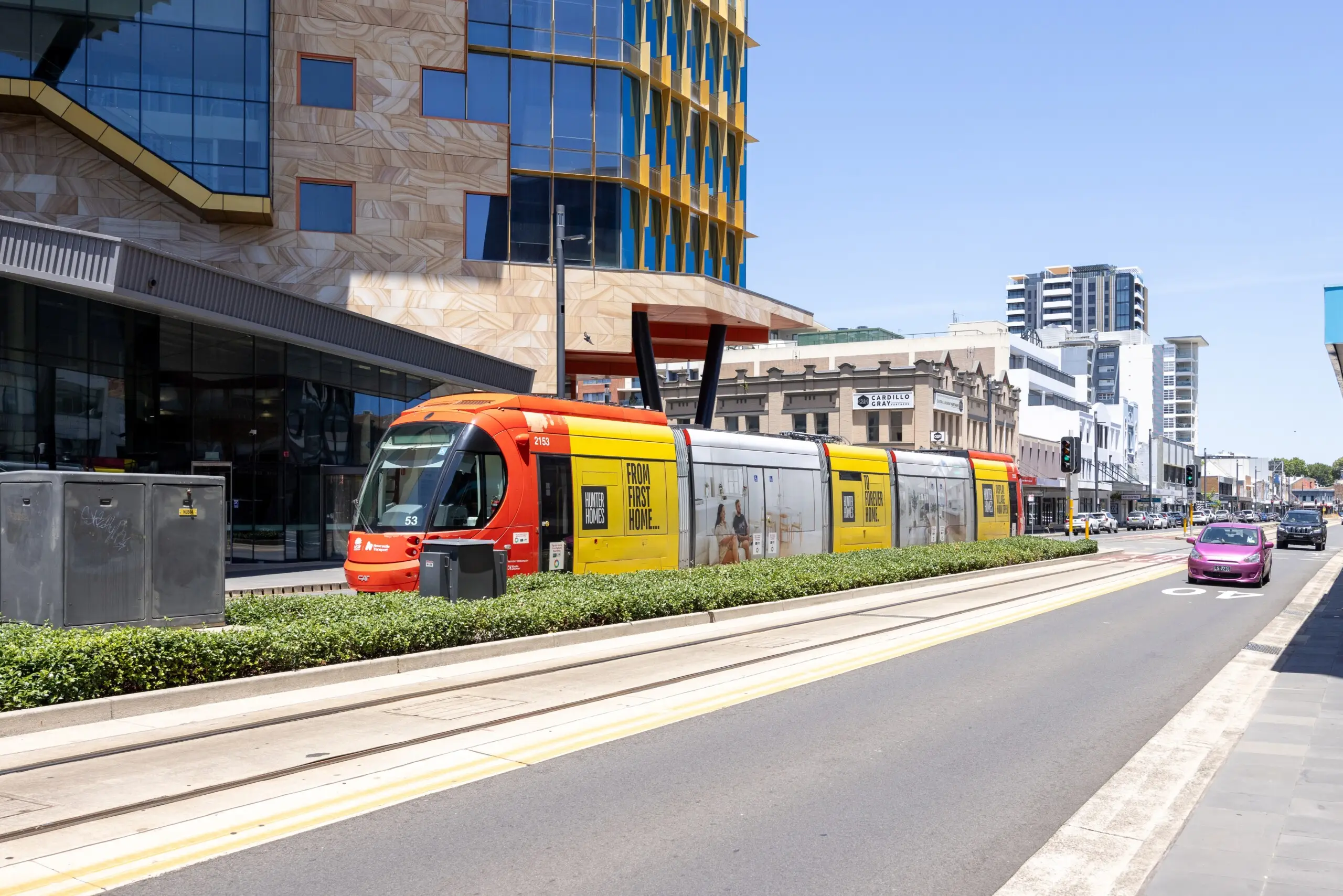 Light rail advertising campaign on a Newcastle tram travelling through the city centre surrounded by modern buildings and busy traffic.