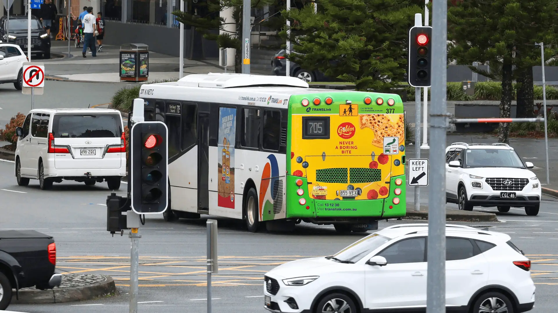 A TransLink public bus with a bright yellow and green advertisement for Golden Bakery crumpets waits at a red light in a busy city intersection. The ad reads, 