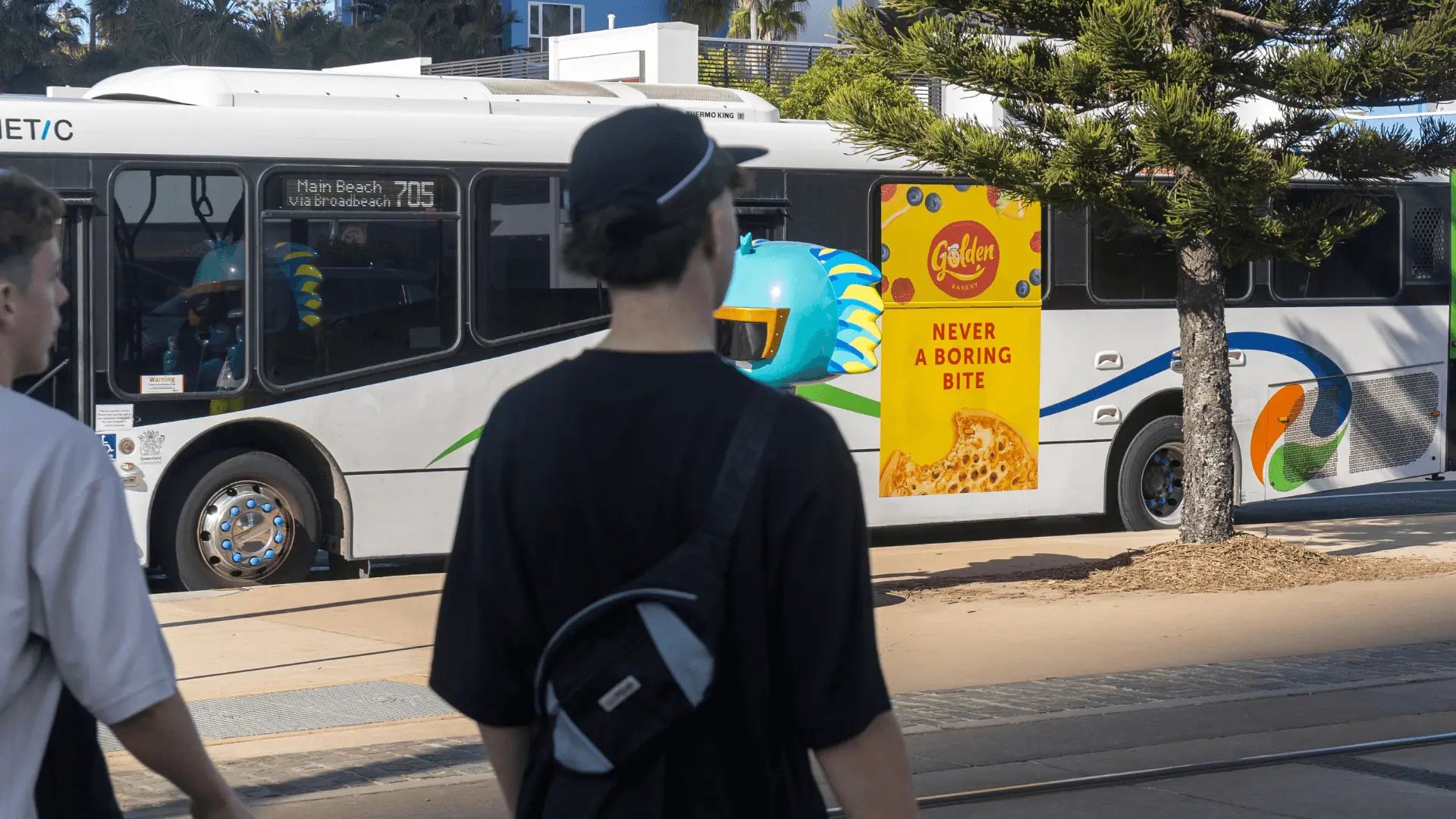 A TransLink bus parked near a tram stop features a bright yellow Golden Bakery ad with the slogan 