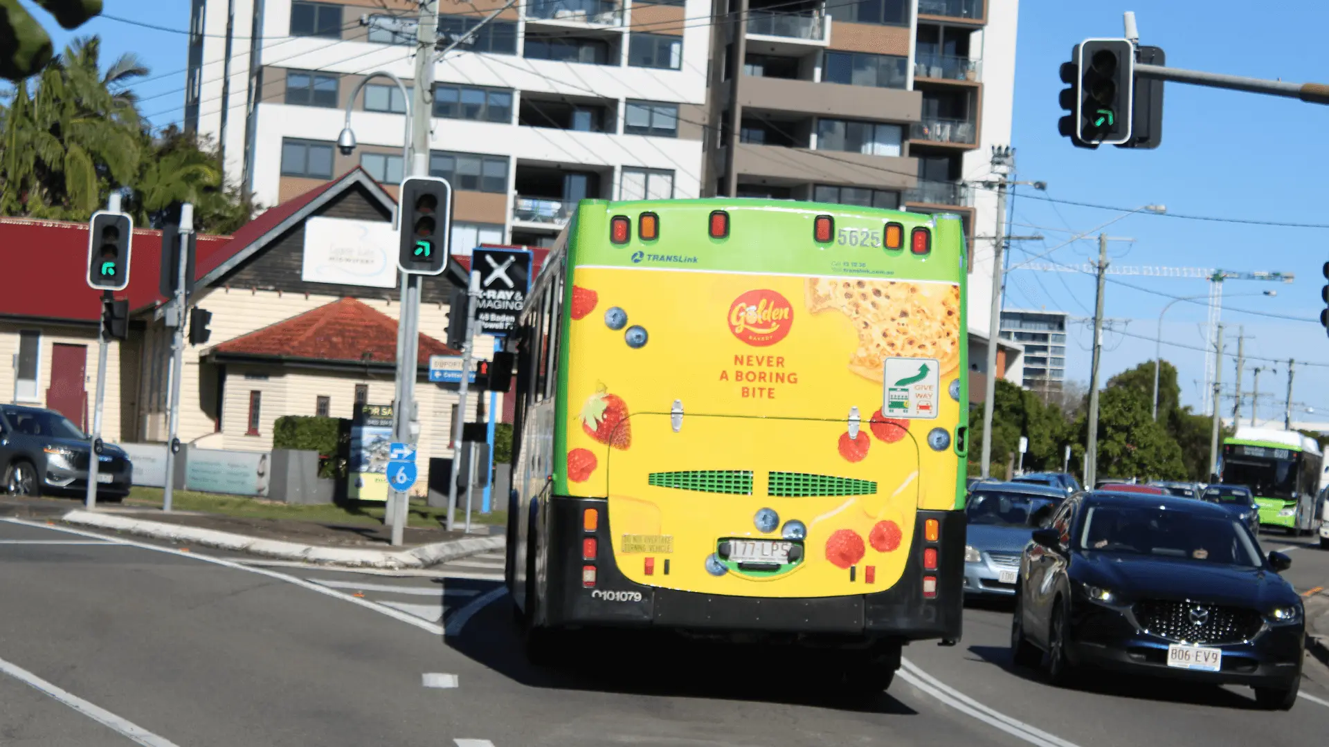 A TransLink bus featuring a bright yellow Golden Bakery crumpet ad drives through an intersection on a sunny day. The ad reads, 