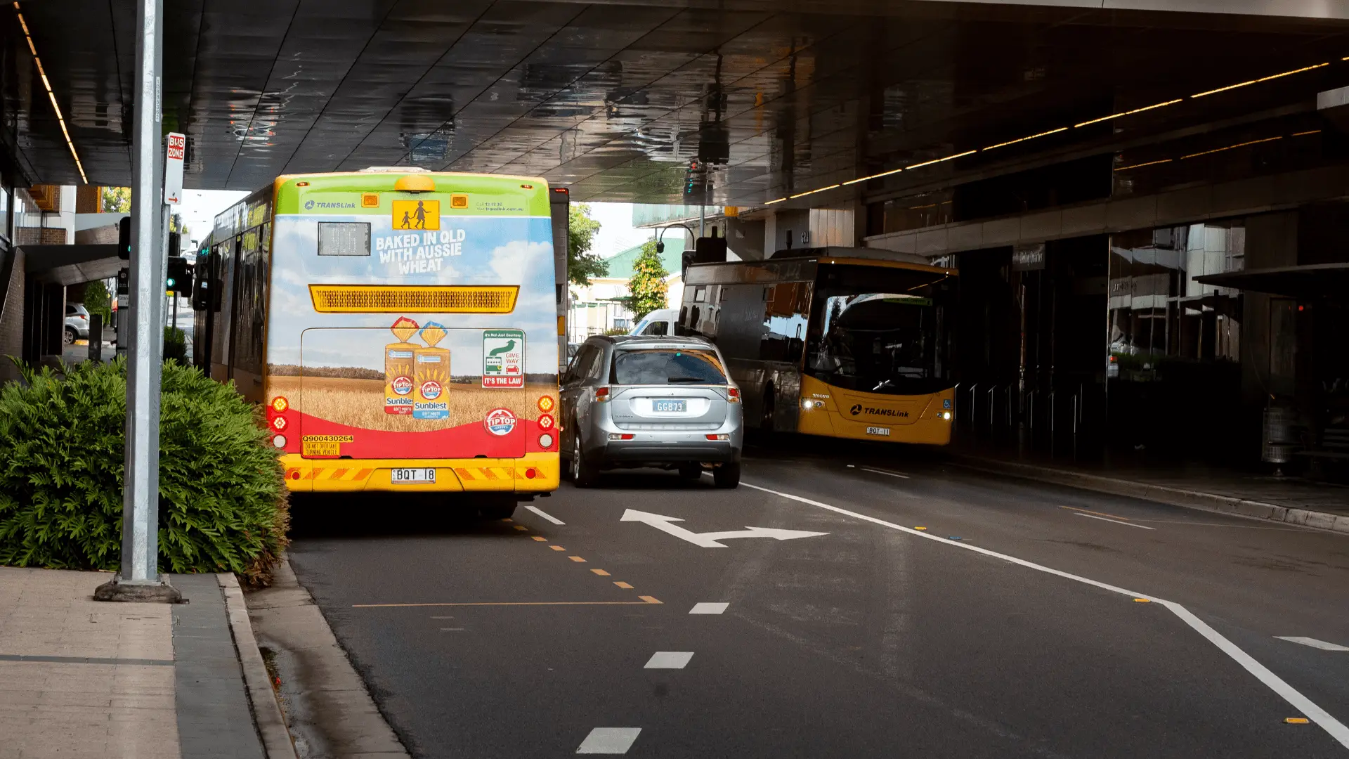 A TransLink bus with a Sunblest bread advertisement drives through a bus zone under a reflective ceiling. The ad features loaves of bread and the text 