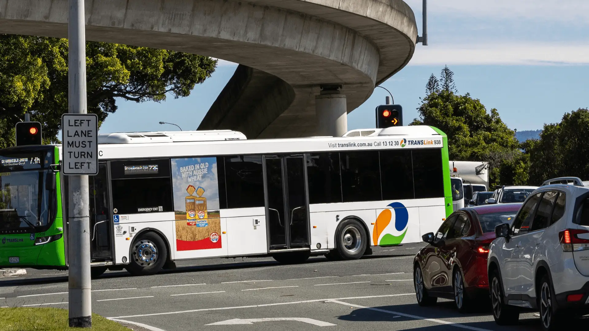 A TransLink bus featuring a Sunblest bread advertisement waits at a red light under an overpass. The ad shows two loaves of bread and reads 