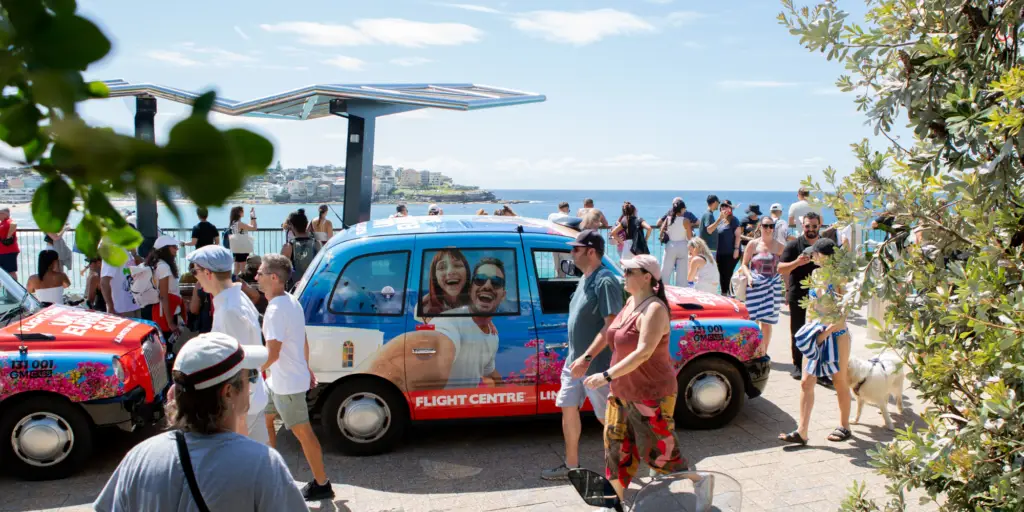 A Flight Centre-branded taxi promoting a "UK & Europe Sale" is parked along a scenic walkway at Bondi Beach. The advertisement features a smiling couple against a vibrant travel-themed backdrop. The area is bustling with beachgoers, some carrying towels, others engaged in conversation, and a dog walking alongside its owner. Lush greenery frames the image, while the blue ocean and coastal landscape stretch into the background.