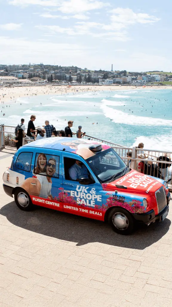 A Flight Centre-branded taxi promoting a "UK & Europe Sale" is parked along a walkway overlooking Bondi Beach. The taxi features a vibrant advertisement with a smiling couple, bright blue and red colors, and the text "Limited Time Deals." The ocean, sandy beach, and city skyline are visible in the background as beachgoers walk along the scenic coastal path.