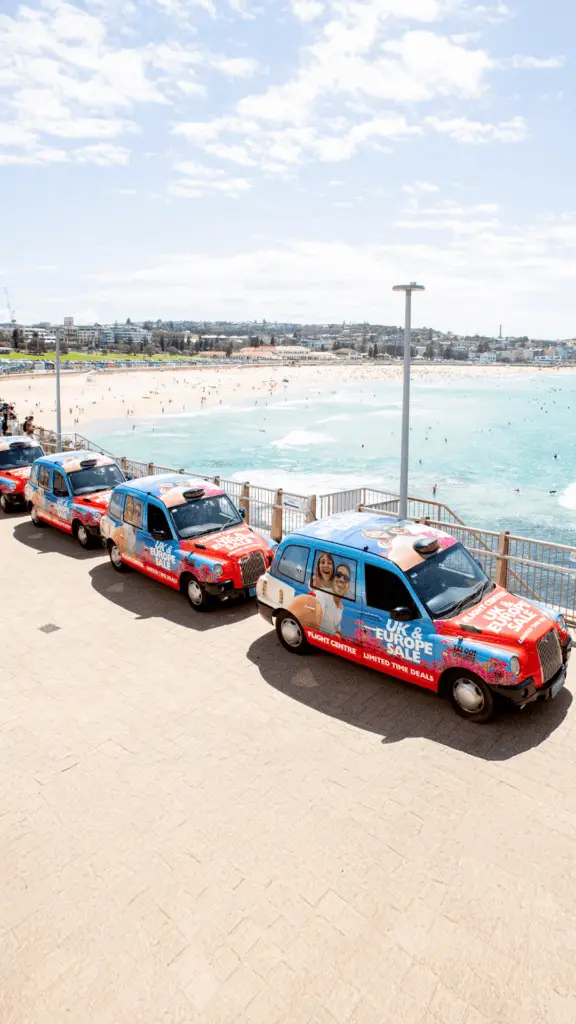A row of classic London-style taxis wrapped in Flight Centre advertisements promoting a "UK & Europe Sale" is parked along a walkway with a view of Bondi Beach. The taxis feature bright red and blue branding with images of smiling travelers. The sandy beach, ocean waves, and distant cityscape create a picturesque backdrop.

