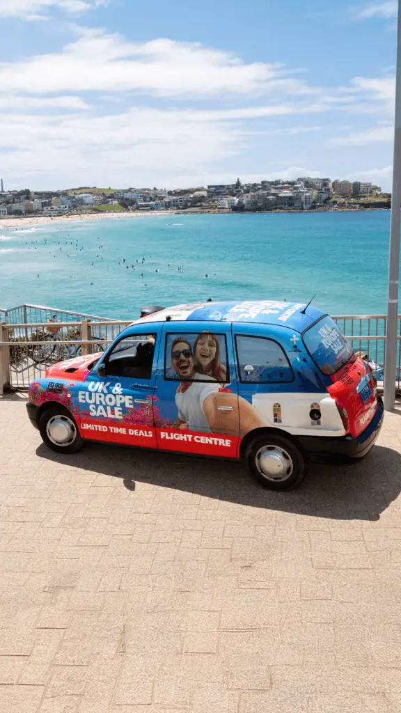 A Flight Centre-wrapped taxi with a "UK & Europe Sale" advertisement is parked by a railing overlooking the blue waters of Bondi Beach. The ad features a smiling couple, white architecture, and pink flowers, evoking a European travel experience. The clear sky and ocean waves complete the vibrant beachside setting.