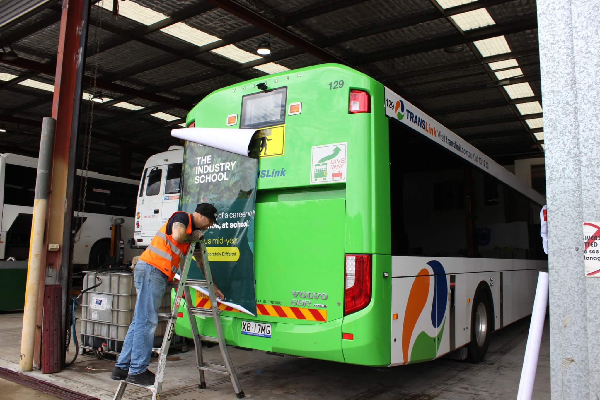 A installer in a hi-vis vest applies an advertisement to the rear of a green TRANSLink bus inside a depot.