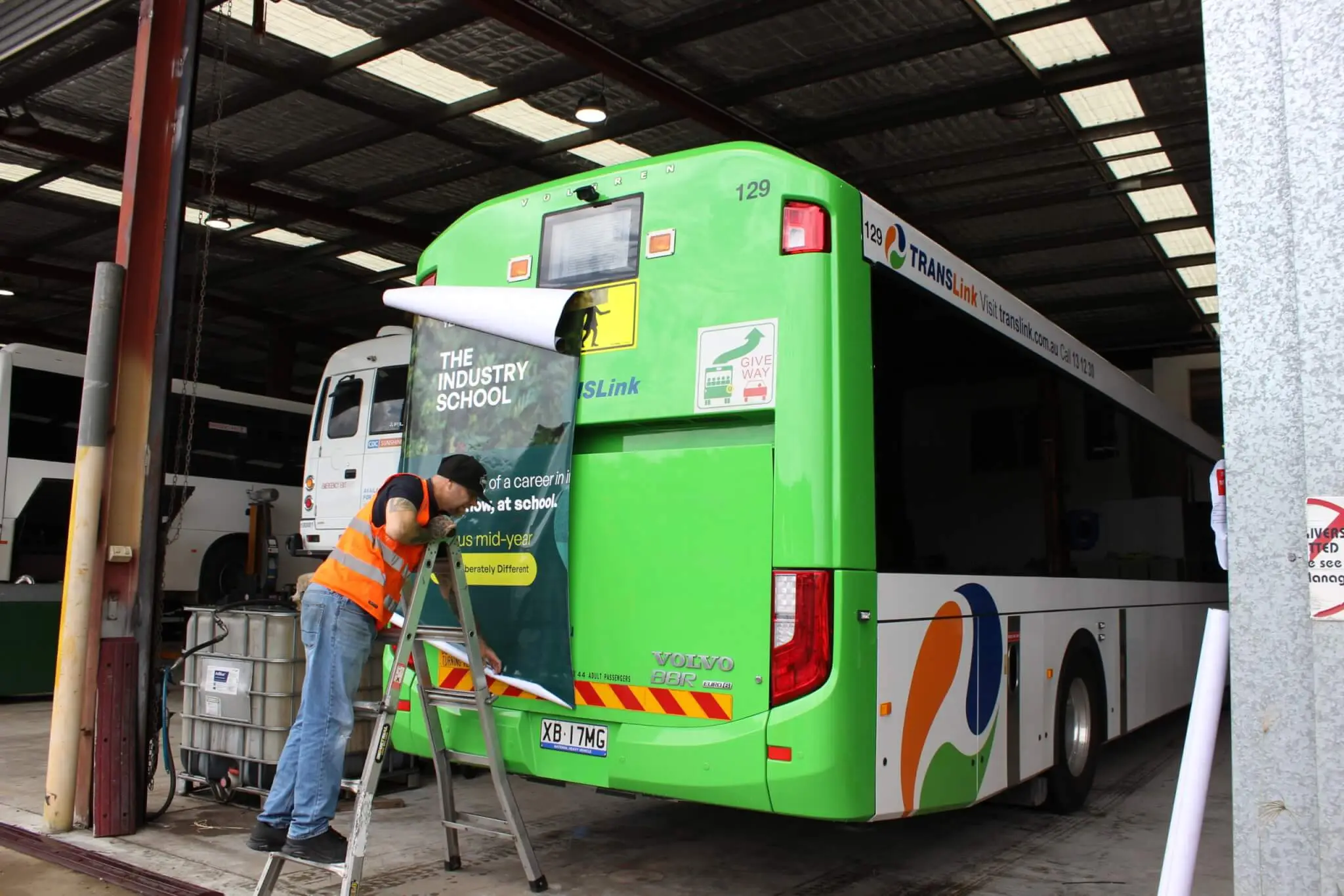 A installer in a hi-vis vest applies an advertisement to the rear of a green TRANSLink bus inside a depot.