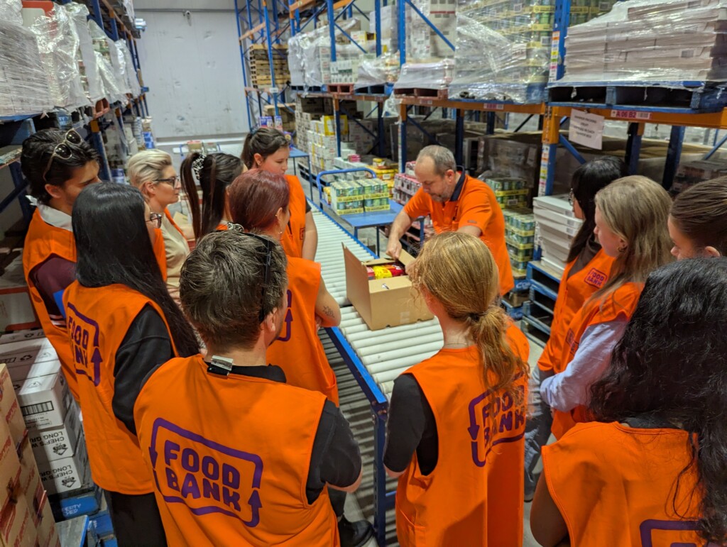 Volunteers in orange Foodbank vests gather around a staff member for packing instructions beside a warehouse conveyor and shelves of donated food.