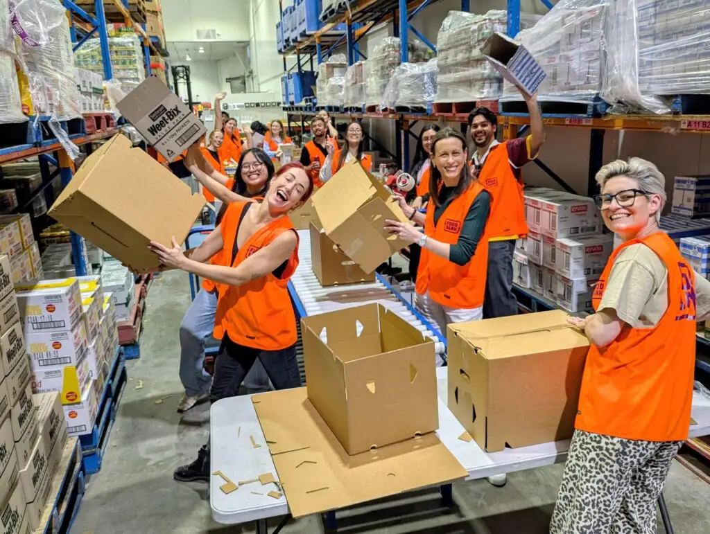 Smiling volunteers in orange vests assemble cardboard boxes at tables between warehouse aisles during the Foodbank Queensland hamper-packing session.