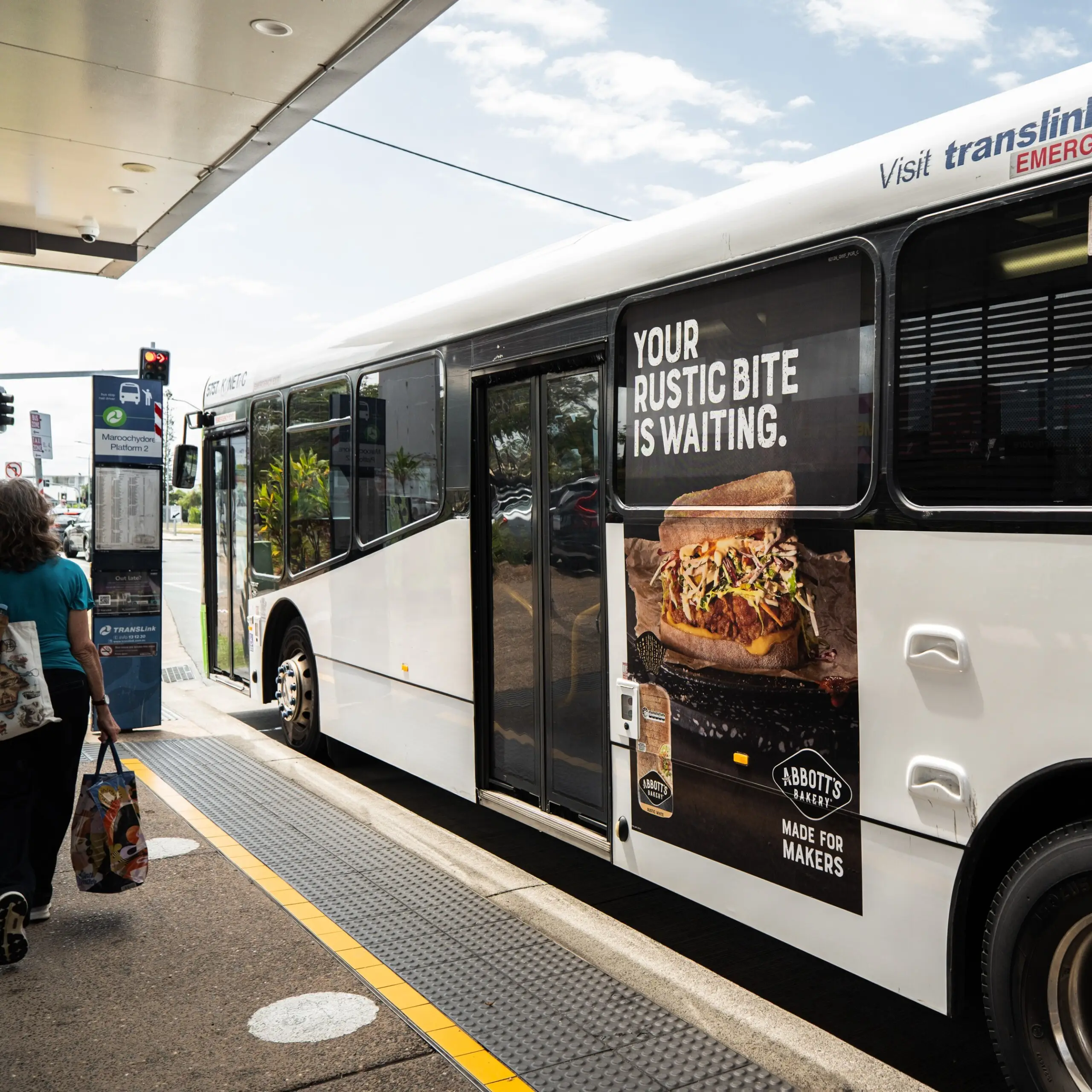 Portrait panel advertisement for Abbott’s Bakery on the side of a TransLink bus at a Maroochydore bus stop, featuring a large burger creative.