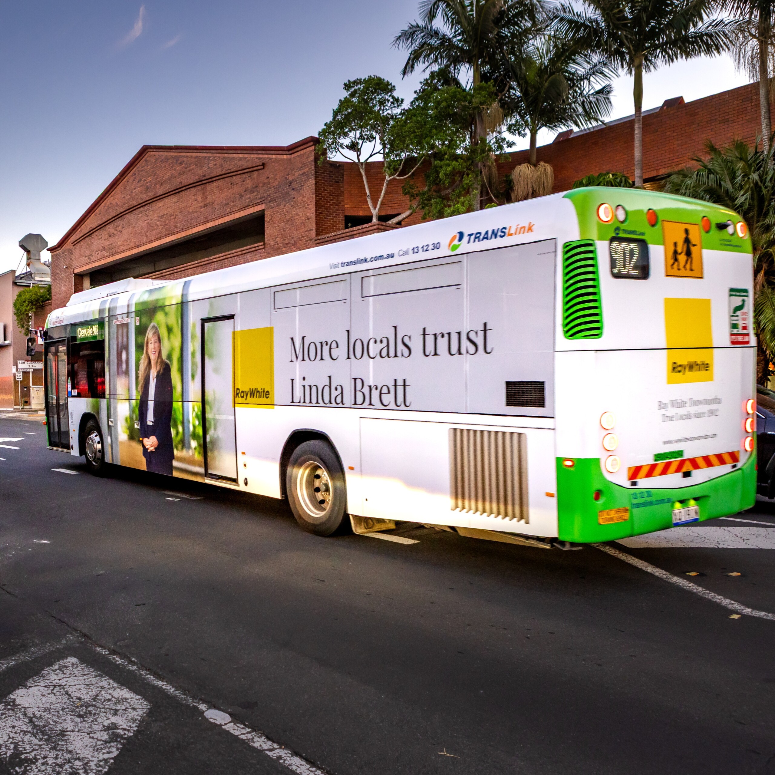 Full bus wrap featuring Ray White real estate agent Linda Brett on a TransLink bus driving through a suburban street at dusk.