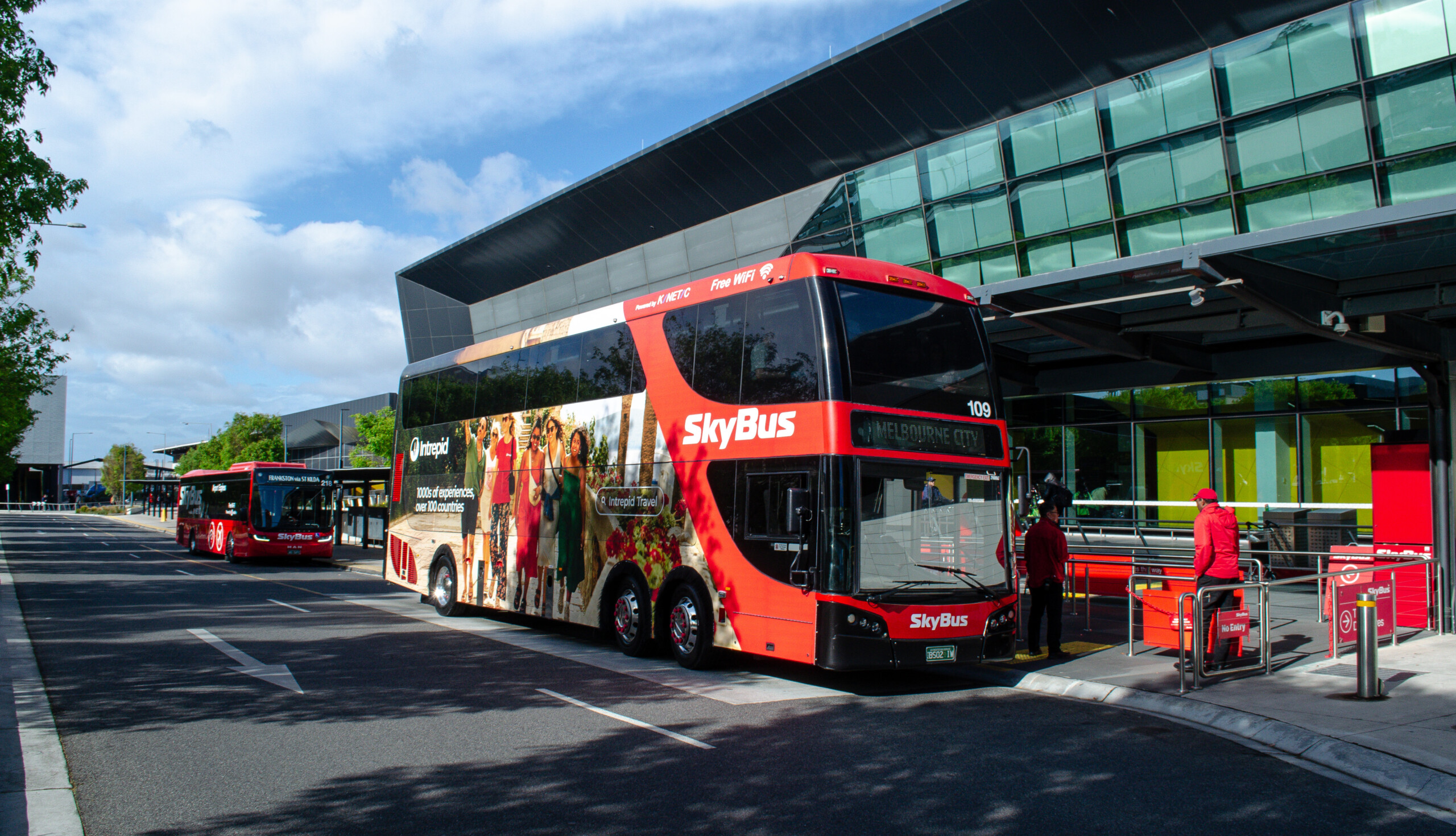 SkyBus advertising featuring a full-side Intrepid Travel campaign on a red double-decker SkyBus at Melbourne Airport with passengers nearby.