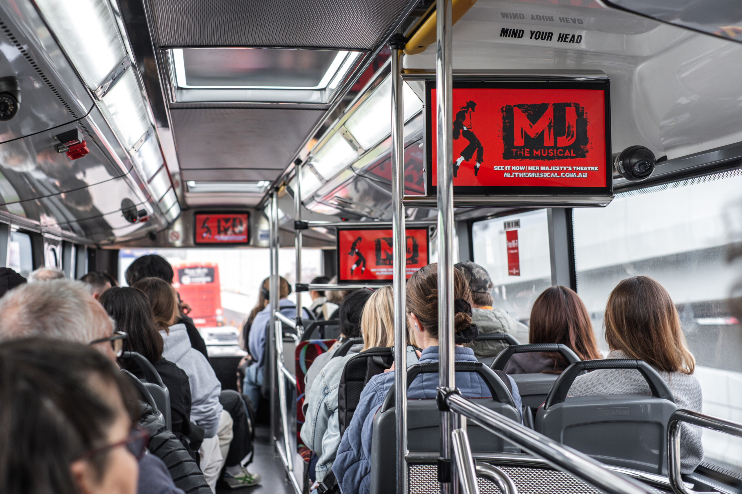 Skybus internal screens displaying MJ The Musical advertising above seated passengers inside a busy SkyBus in Melbourne.