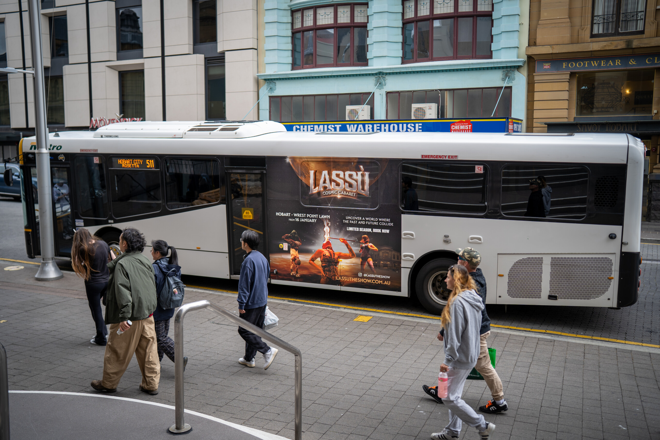 City bus featuring transit advertising panel as pedestrians walk past in Hobart city centre.