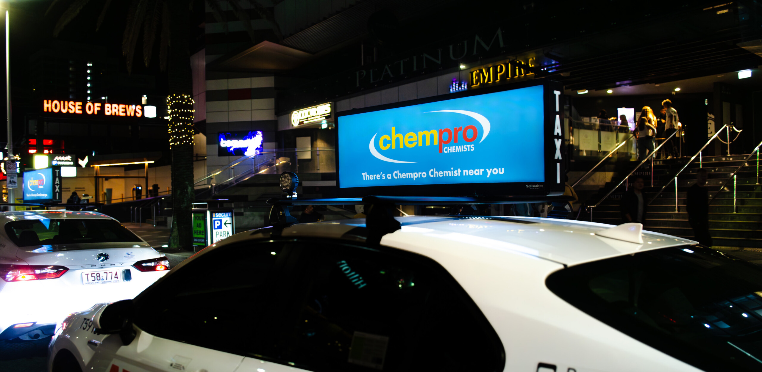 Static taxi tops advertising featuring a Chempro Chemists campaign on a taxi travelling through a busy nightlife entertainment precinct.